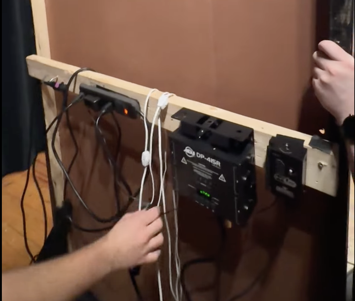 The rear of the flat that the On Air sign is on. A 2x4 drilled onto the back of the flat has various hardware on it. From left to right, it has a power strip, a DMX dimmer, and a wDMX Rx. Wires that lightbulbs screw into are draped over the 2x4.