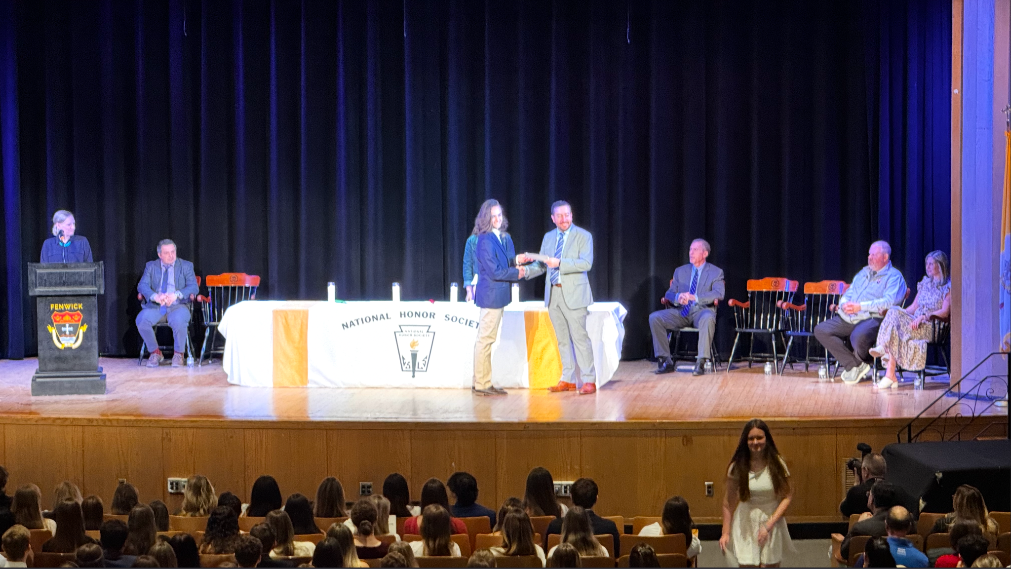 A NHS Induction Ceremony. A speaker stands at a podium on SR, a table is at C, Bishop Fenwick High School Principal Chris Canniff stands near C, shaking an NHS inductee's hand. At both SL and SR, Fenwick administration staff sits in chairs.