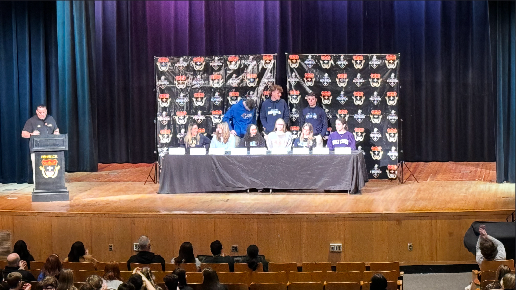 A college athletics signing banquet. A speaker stands at a podium on SR, and at C, there is a long table in front of a Fenwick backdrop with nine students.